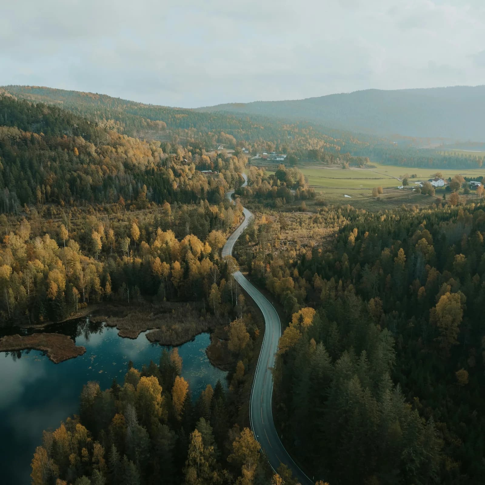 En svingete vei mellom skog i høstfarger, jorder og et lite vann
